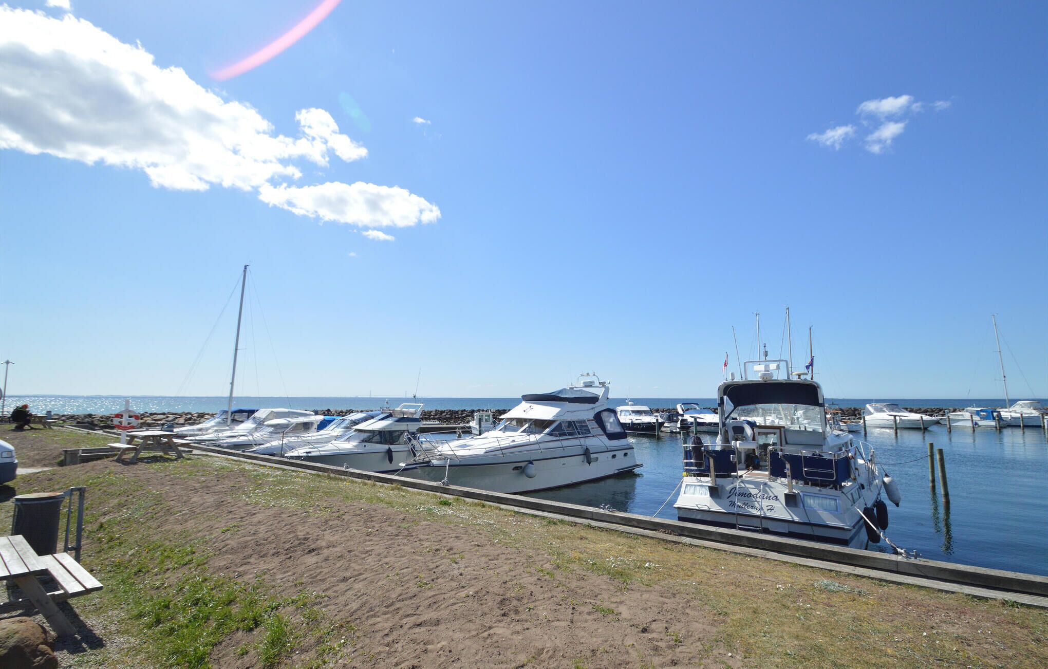 Un port de plaisance où sont amarrés plusieurs bateaux, une zone herbeuse avec des tables de pique-nique et un ciel bleu dégagé.