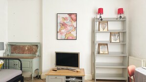Una habitación con una cama, un escritorio con una computadora, un televisor, una estantería y un cuadro en la pared.