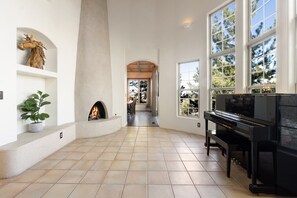 A spacious living area with a fireplace, a piano, and large windows offering a view of trees. - Serenity Skyline Lodge (Colorado Springs)