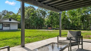 A covered outdoor area with a glass table and chairs, a concrete floor, and a view of a house and trees in the background. - Updated 4 bedroom home in Oragnefield (Orange)