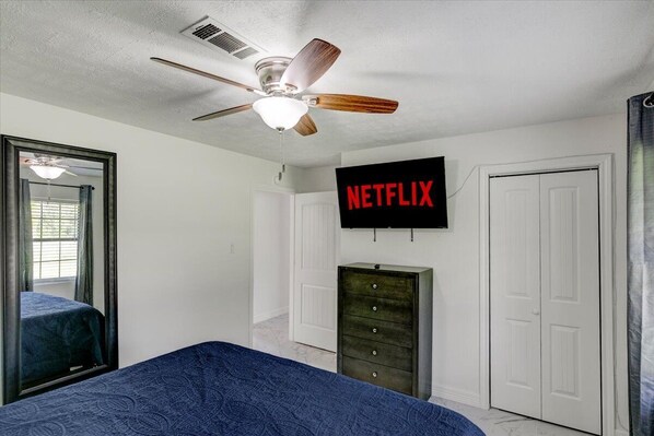 A bedroom with a ceiling fan, a bed with a blue bedspread, a dresser, a television mounted on the wall, and a door.