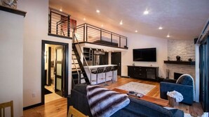 Una sala de estar moderna con un sofá, una mesa de centro y un televisor. Se ve una escalera que sube a un altillo con un escritorio y una silla.