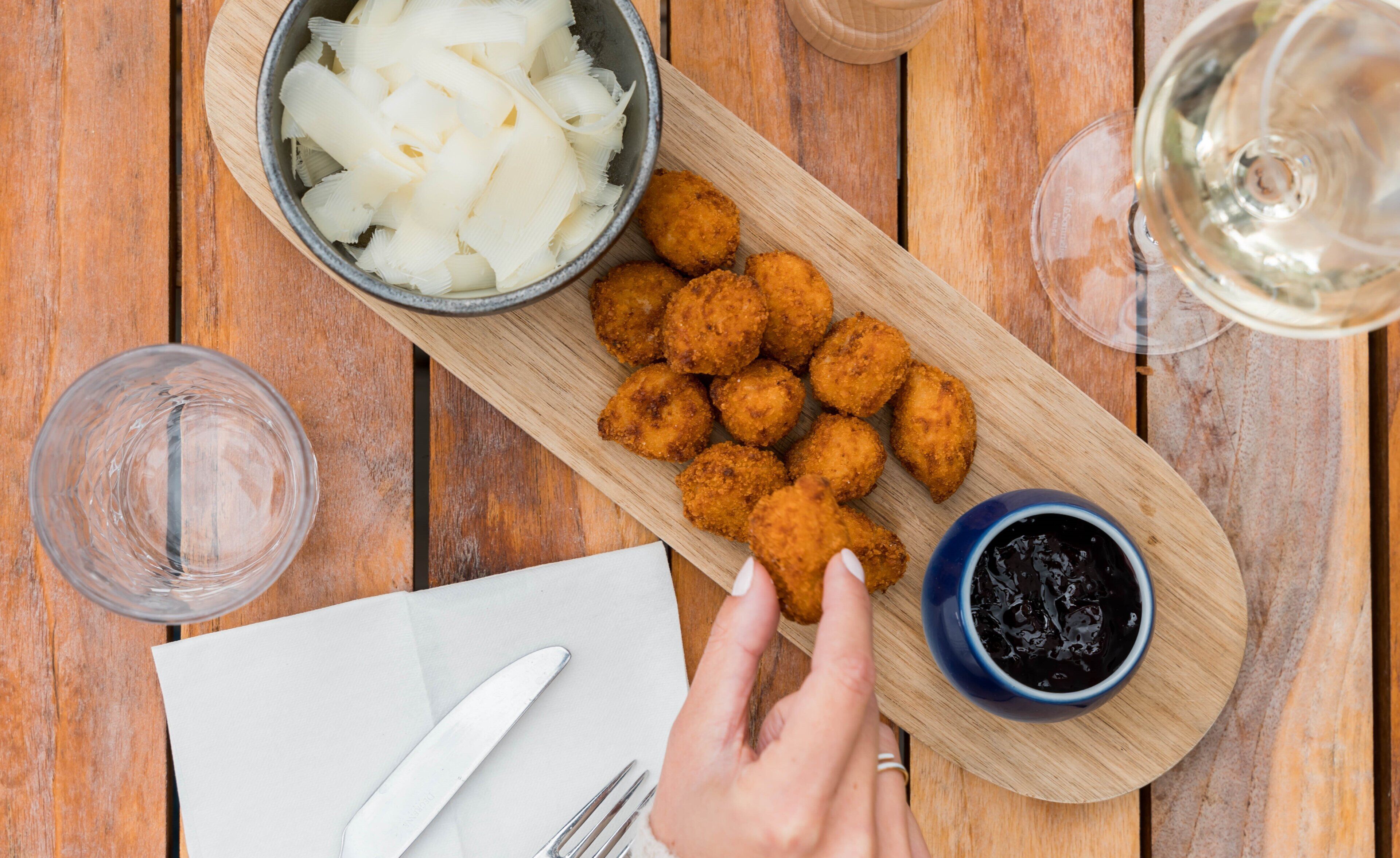 A wooden table with a plate of fried food, a bowl of onions, a bowl of sauce, and a glass of white wine.