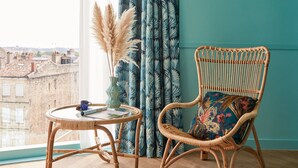 A room with a wicker chair, a small round table, and a view of buildings through a window. - Hotel Indigo Bordeaux Centre Chartrons by IHG (Bordeaux)