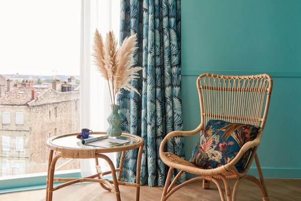 Una habitación con una silla de mimbre, una mesita redonda y vistas de edificios a través de una ventana.
