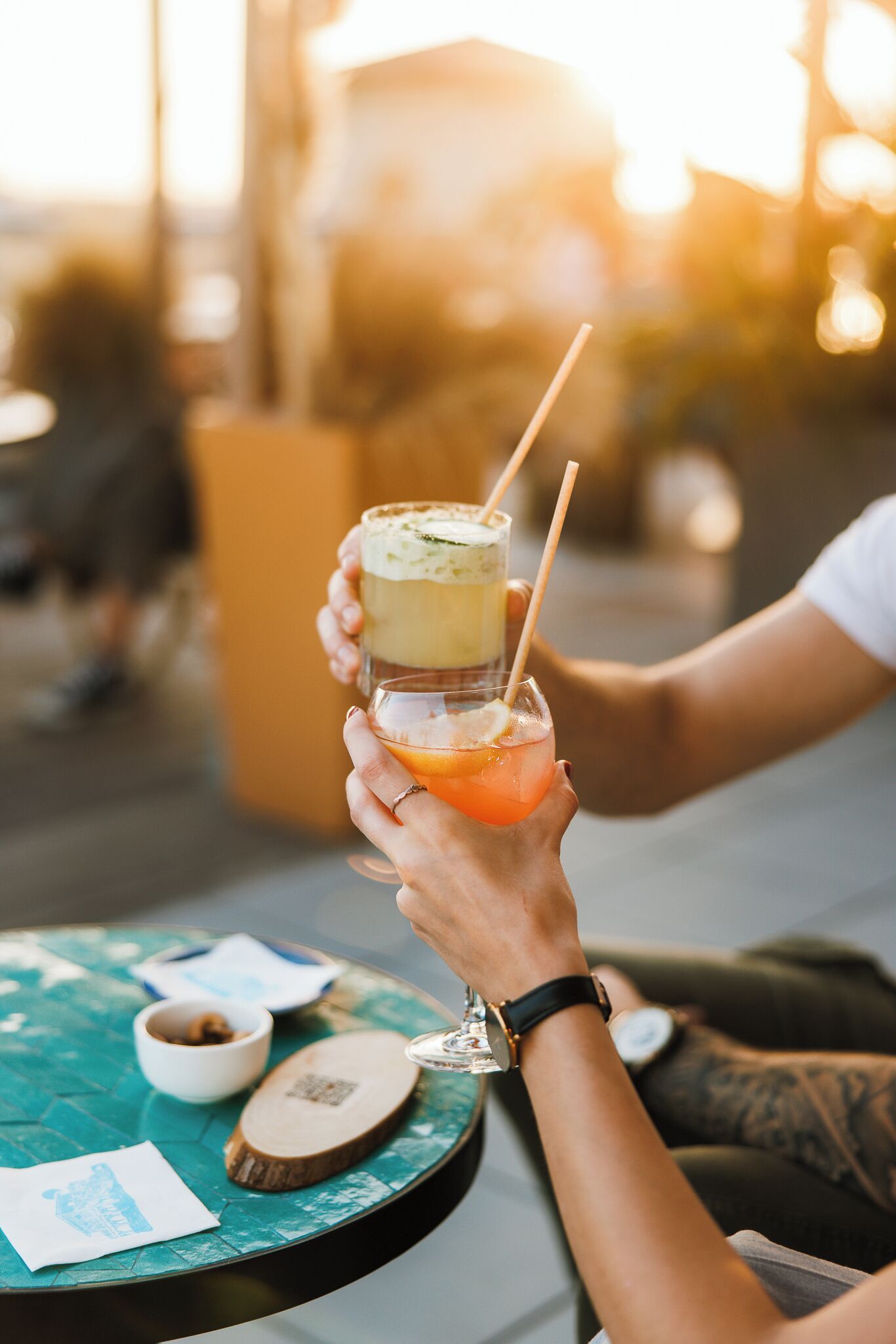 Une personne tenant un verre contenant une boisson à plusieurs couches, assise à une table ronde avec un petit bol et un sous-verre en bois.