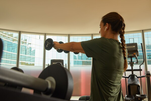 Una persona haciendo ejercicio con mancuernas en un gimnasio con amplias ventanas y equipamiento de entrenamiento.