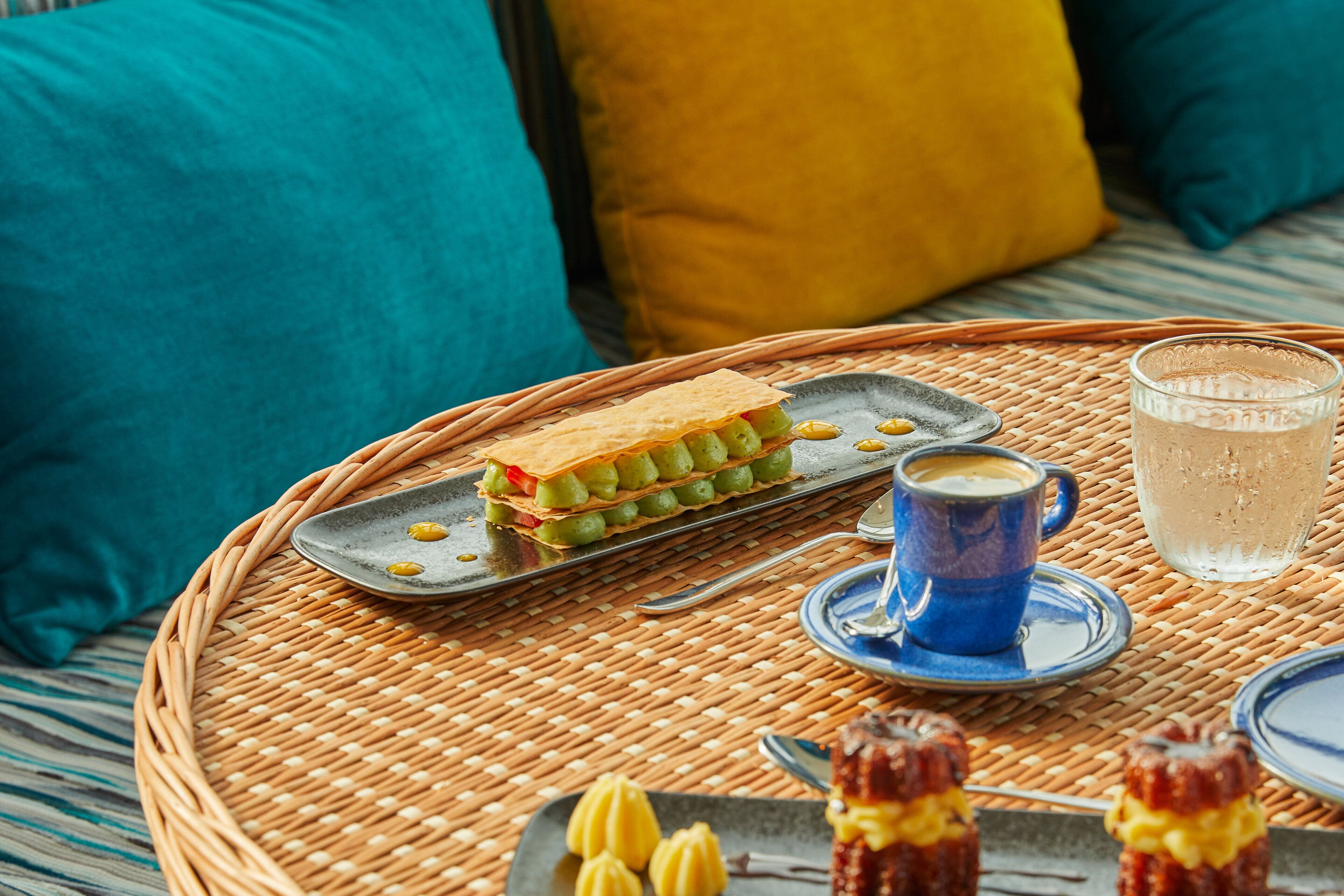 A wicker tray with a sandwich, a glass of water, and a cup of coffee on a patterned tablecloth.
