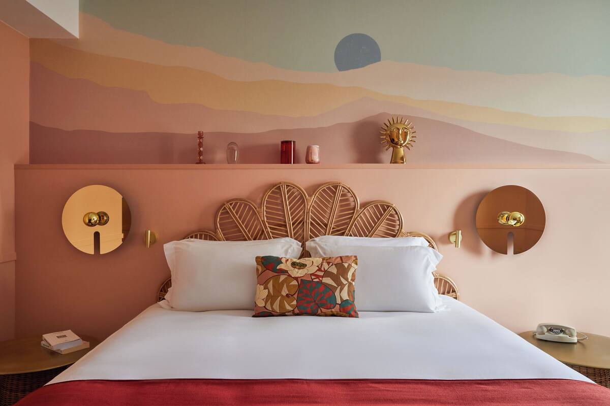 Ein Bett mit weißen Leinen, einem floralen Kissen und einer roten Tagesdecke. Auf einem Nachttisch liegen ein Telefon und ein Buch. Im Raum befindet sich ein wandmontierter Spiegel und ein dekoratives, sonnenähnliches Objekt.