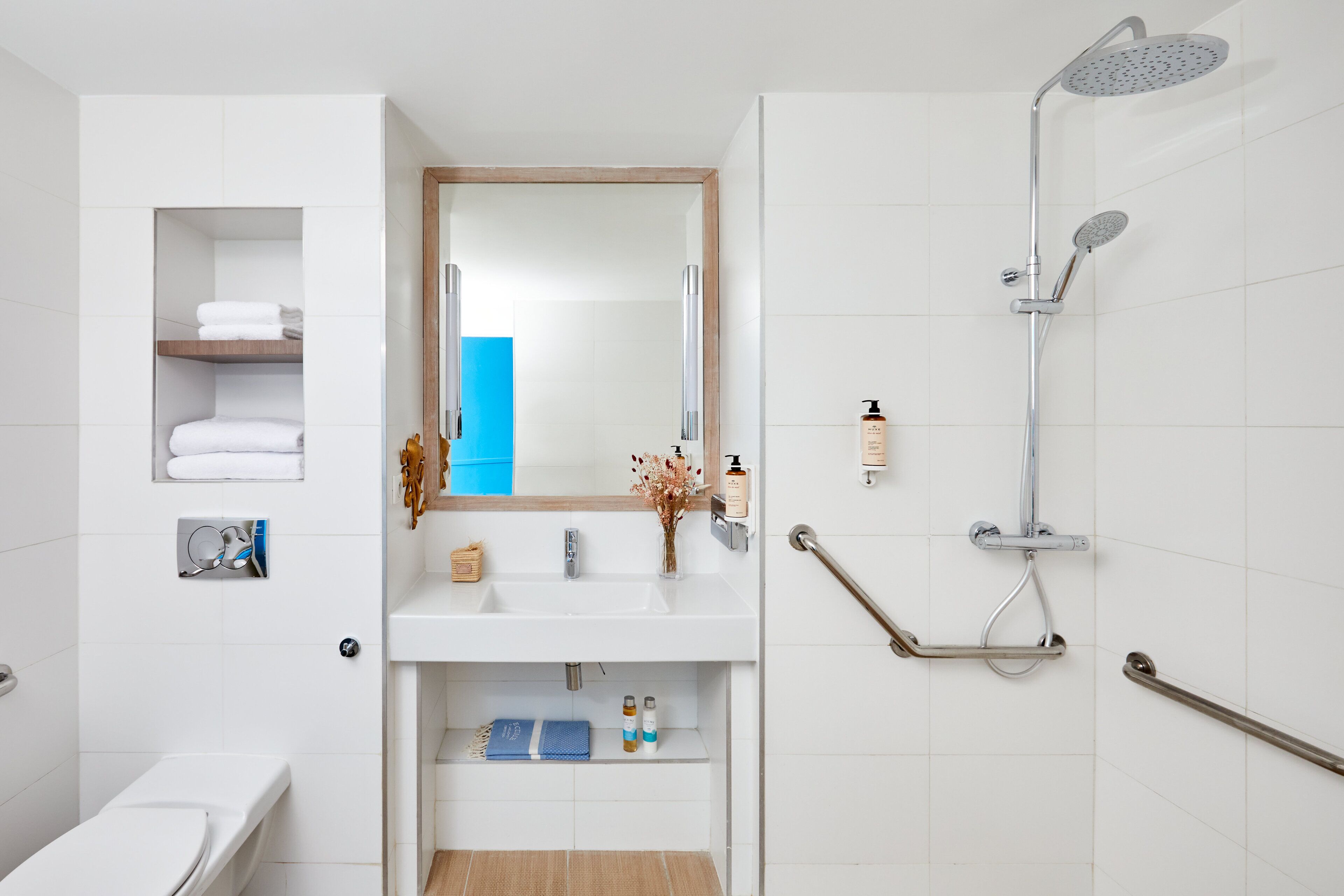 Une salle de bain moderne avec un lavabo blanc, un miroir et une douche. Des barres d’appui sont présentes pour faciliter l’accès, et une serviette bleue est visible.