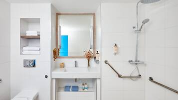 Une salle de bain moderne avec un lavabo blanc, un miroir et une douche. Des barres d’appui sont présentes pour faciliter l’accès, et une serviette bleue est visible.