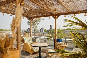 Terraza en la azotea con mobiliario de mimbre, techo de paja y vistas de edificios de la ciudad.