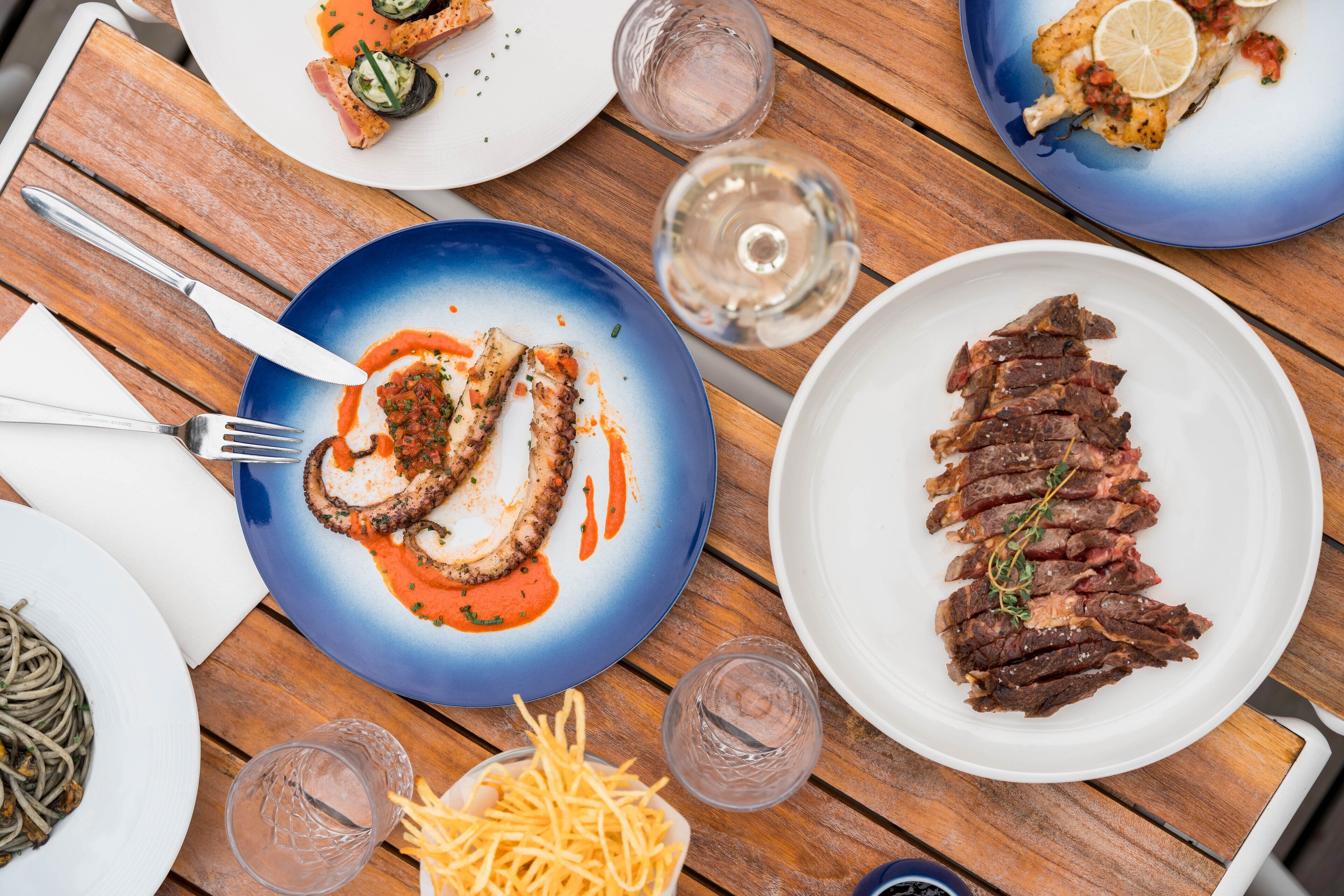 Une table en bois sur laquelle on trouve différents plats, notamment une assiette de steak, une assiette de poulpe et une assiette de frites.