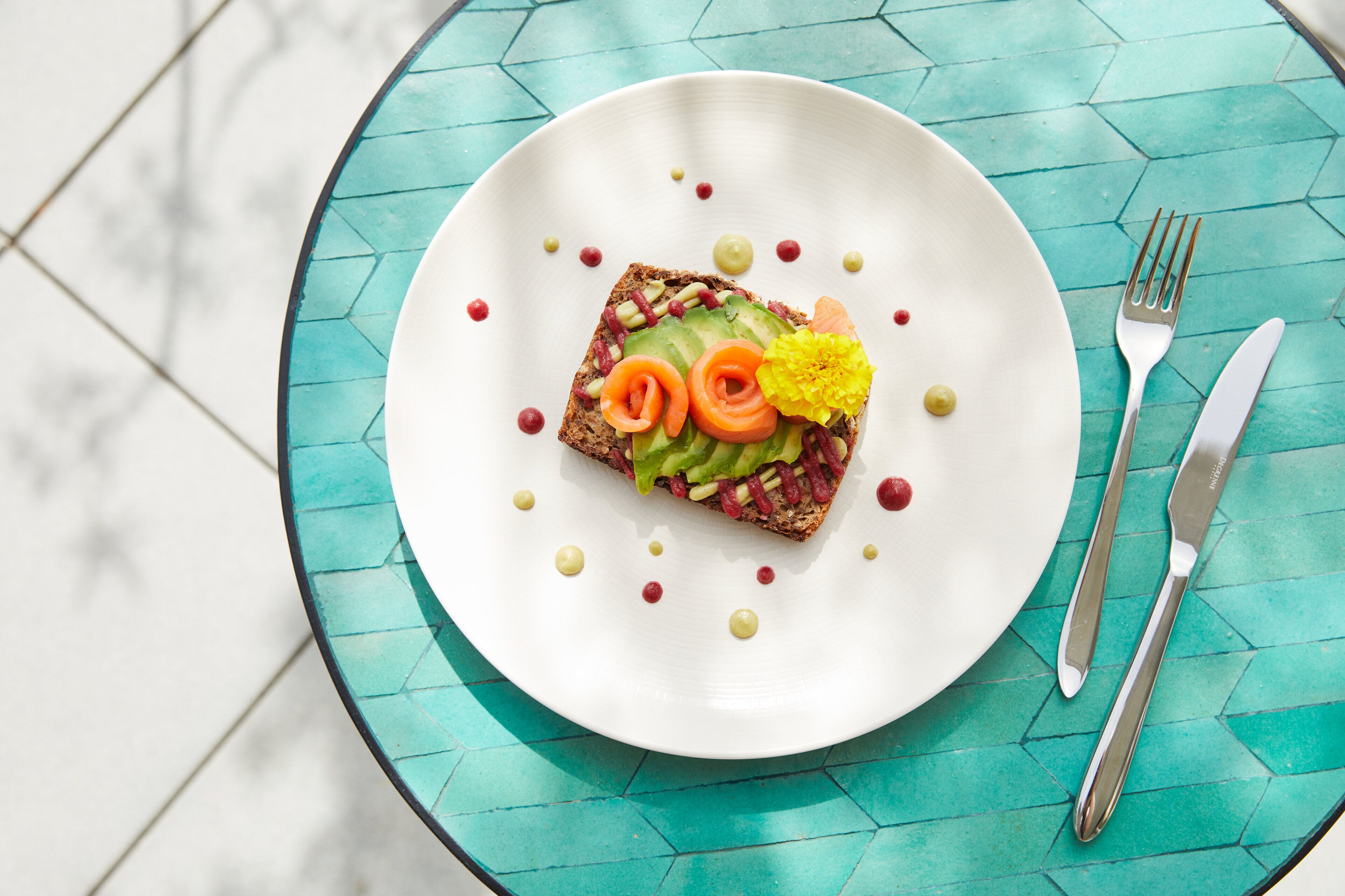 Ein weißer Teller mit Toast, belegt mit Avocado, Tomate und einer Blüte, auf einem türkisfarbenen Tisch.