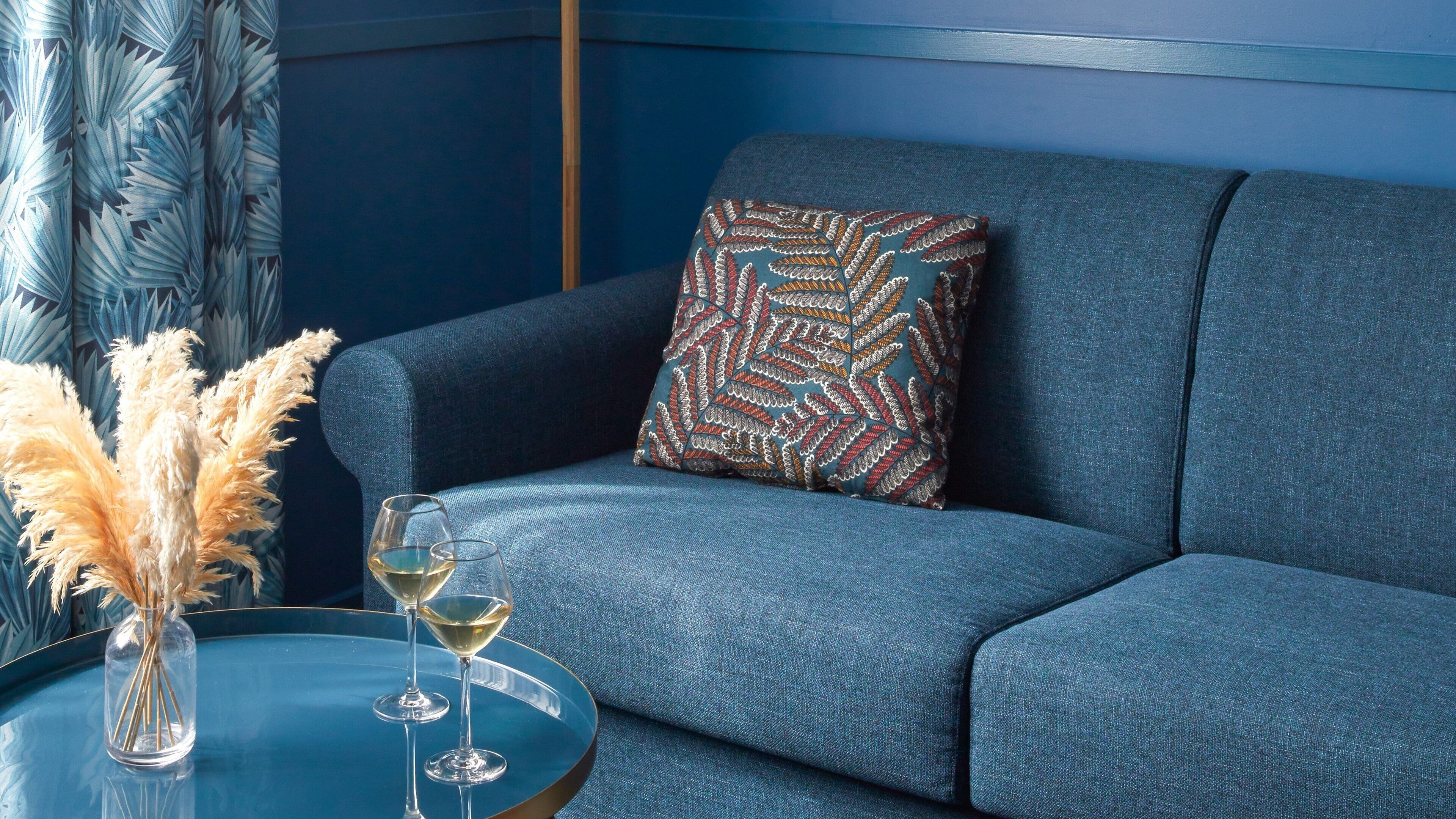 A blue sofa with a patterned pillow, a glass table with two wine glasses and a vase of dried pampas grass.