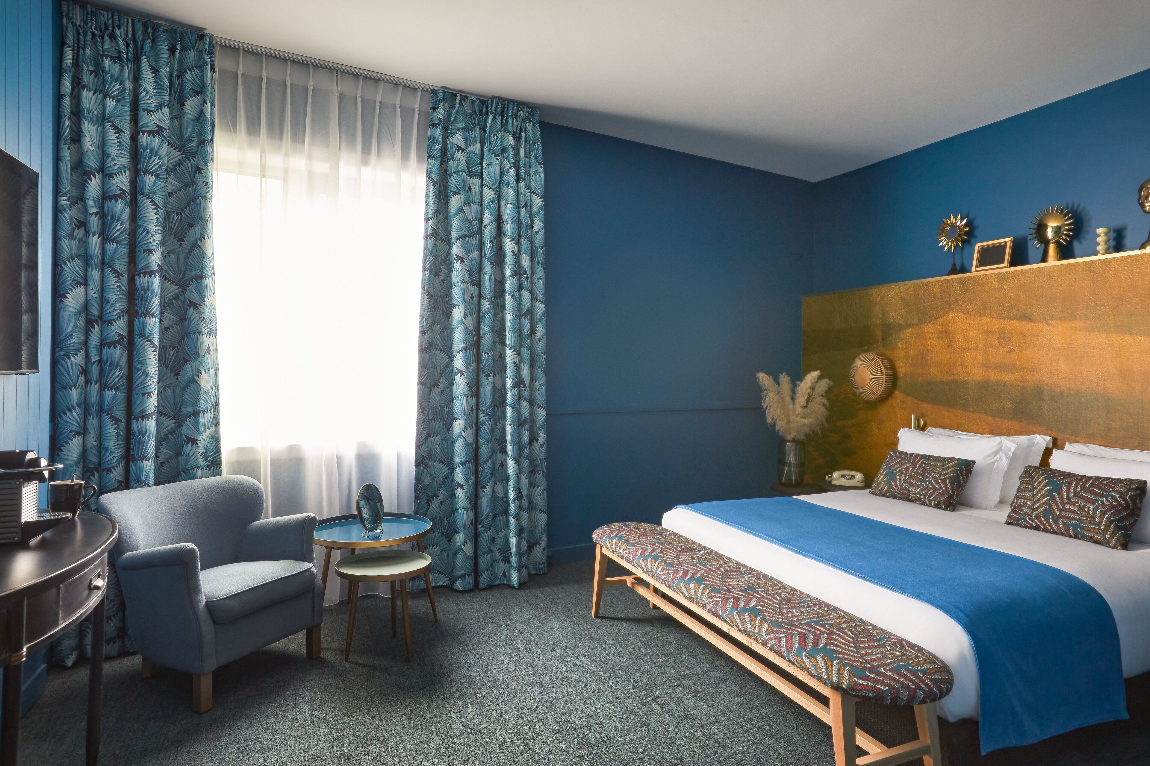 A hotel room with a bed, a desk, a chair, a small table, and a bench.
