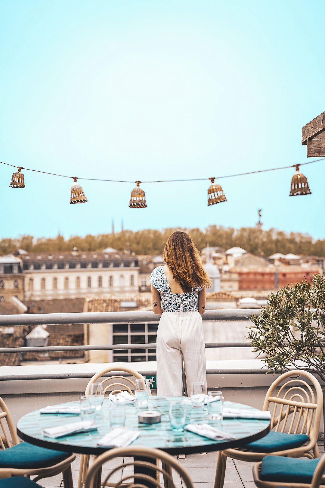 Eine Frau steht auf einem Balkon mit Blick auf eine Stadtlandschaft, mit Lichterketten und einem Essplatz.