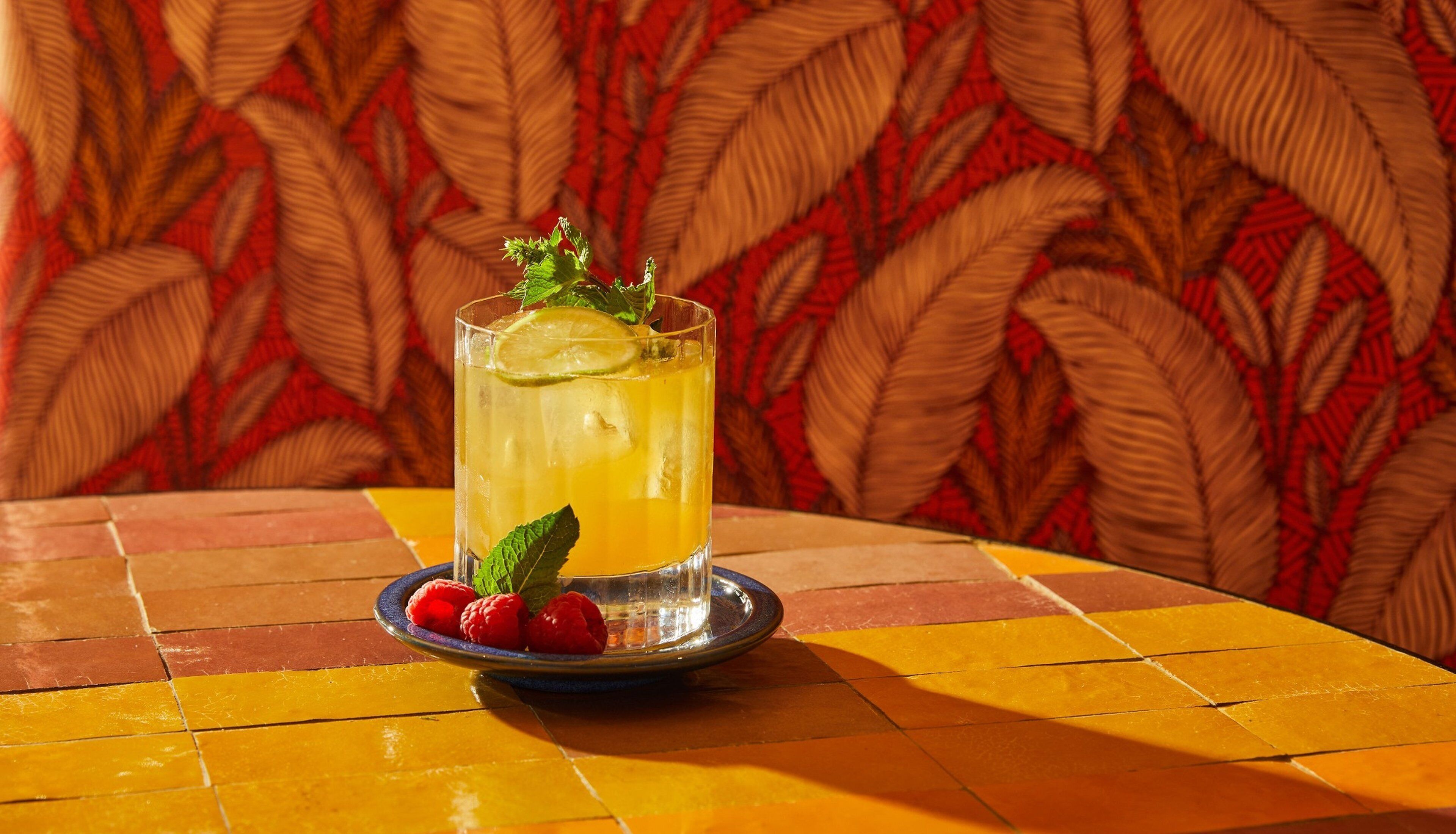 A glass of yellow beverage with a lime slice and mint garnish on a plate with raspberries, set on a tiled table with a patterned backdrop.