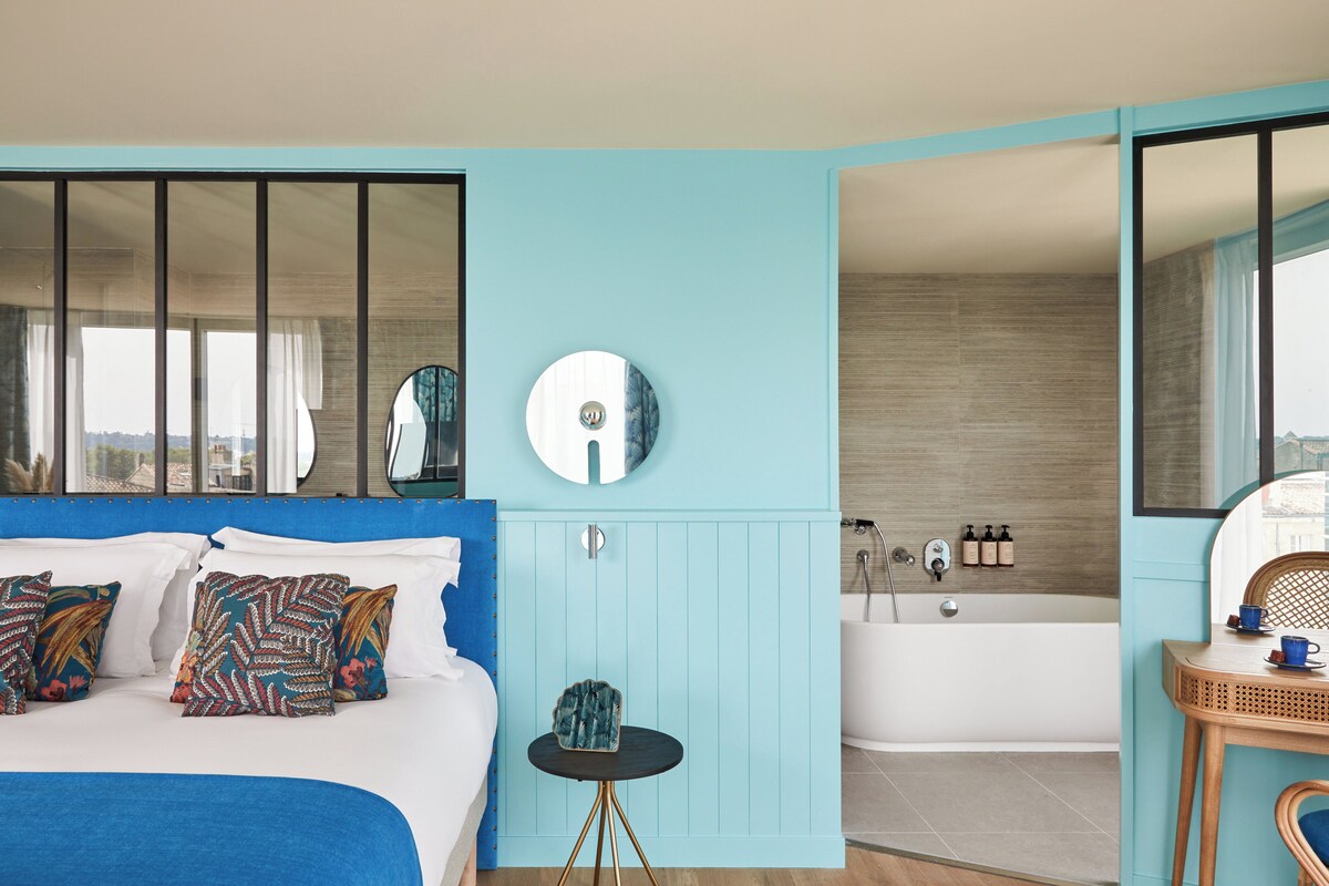 Een moderne slaapkamer met een groot bed, een ronde spiegel, een klein tafeltje en een badkamer met bad en douche.