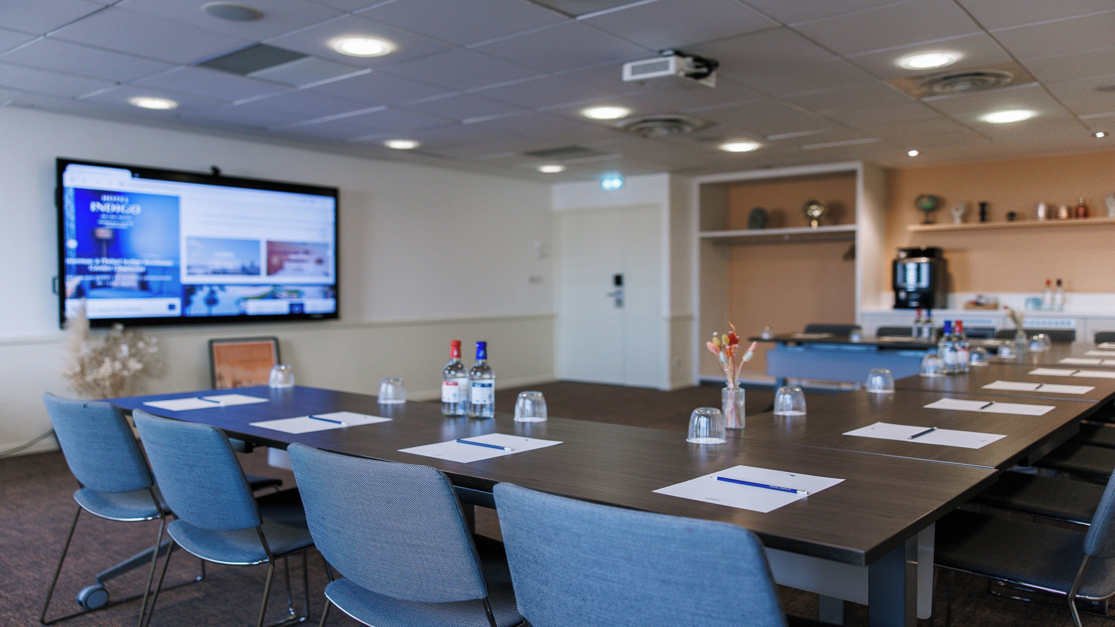 A conference room with a long table, chairs, a projector screen, and a kitchenette area.
