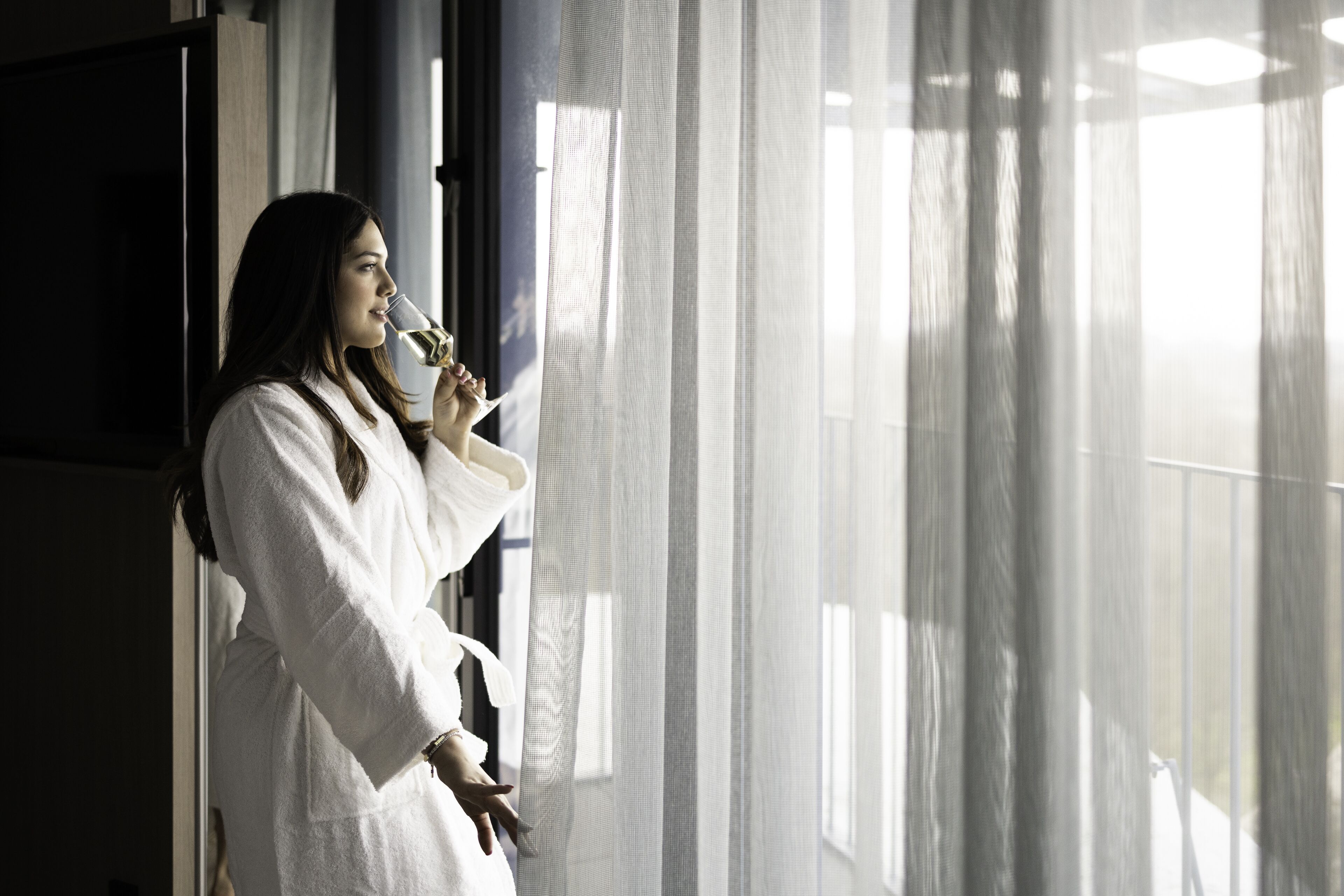 Una persona vestida con una bata blanca está de pie junto a una ventana con cortinas traslúcidas, un televisor montado en la pared y una puerta a la vista.