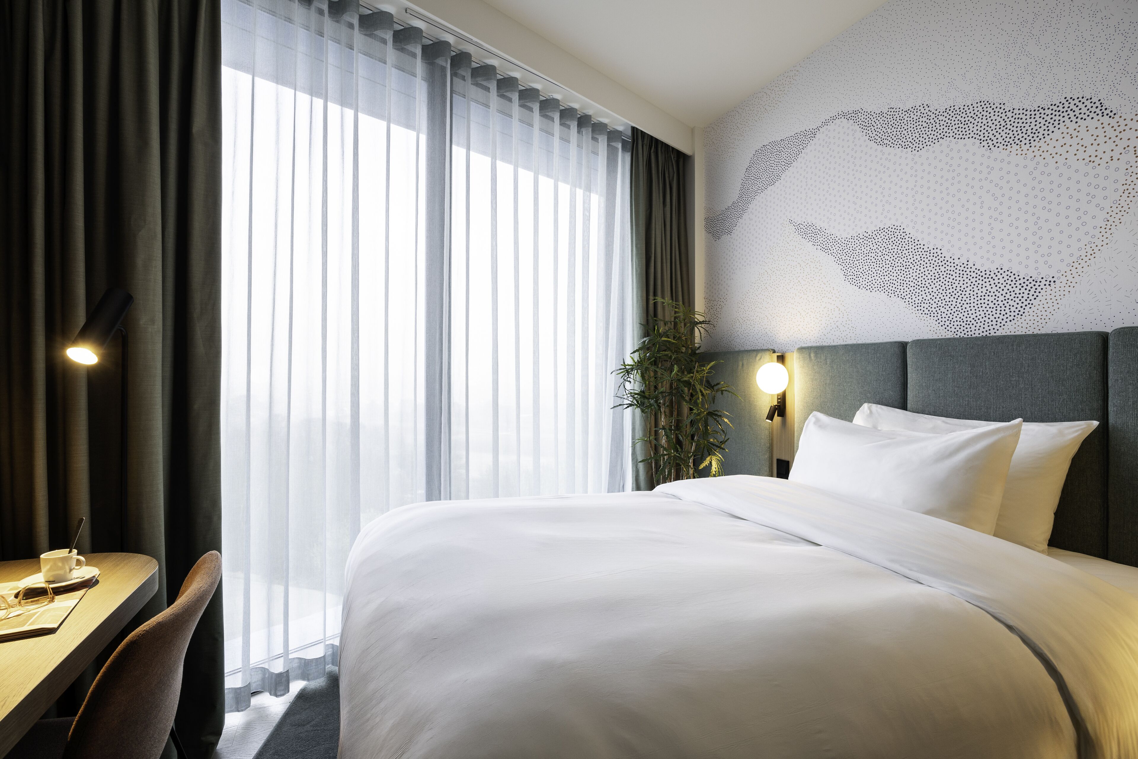 Habitació d'hotel amb un llit gran, un escriptori amb una cadira, una làmpada i una planta.