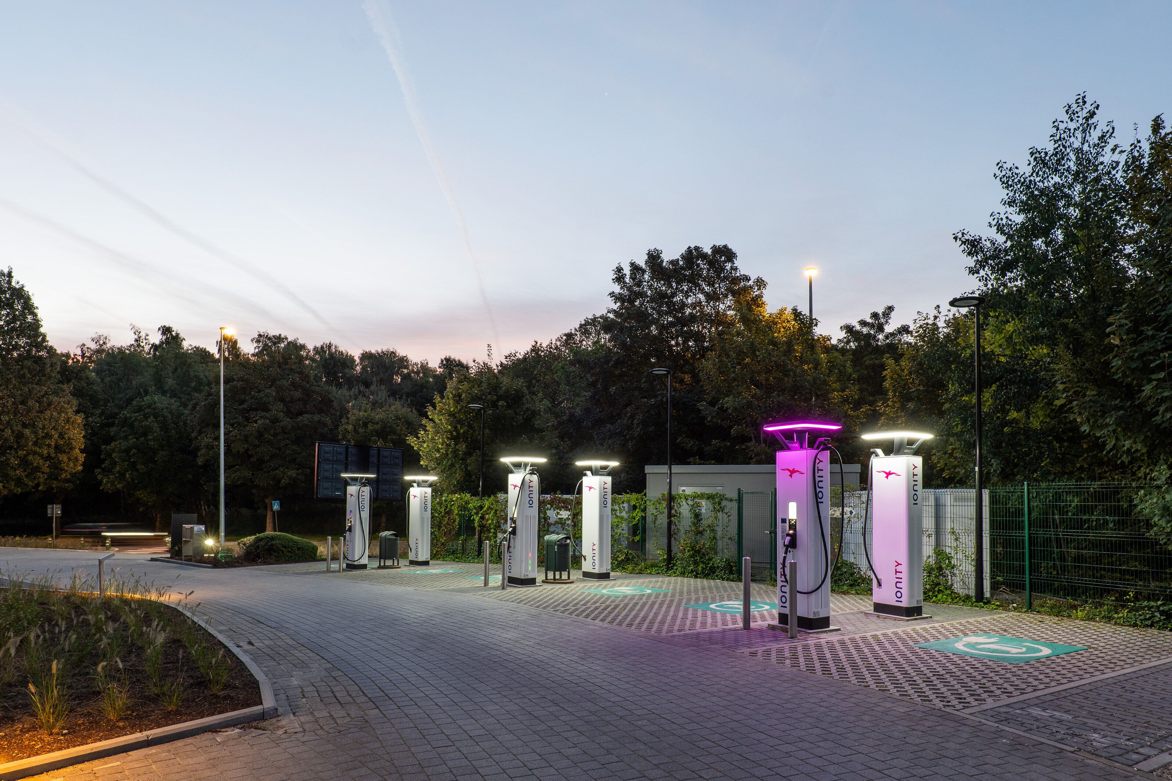 A row of electric vehicle charging stations with a paved walkway and trees in the background.