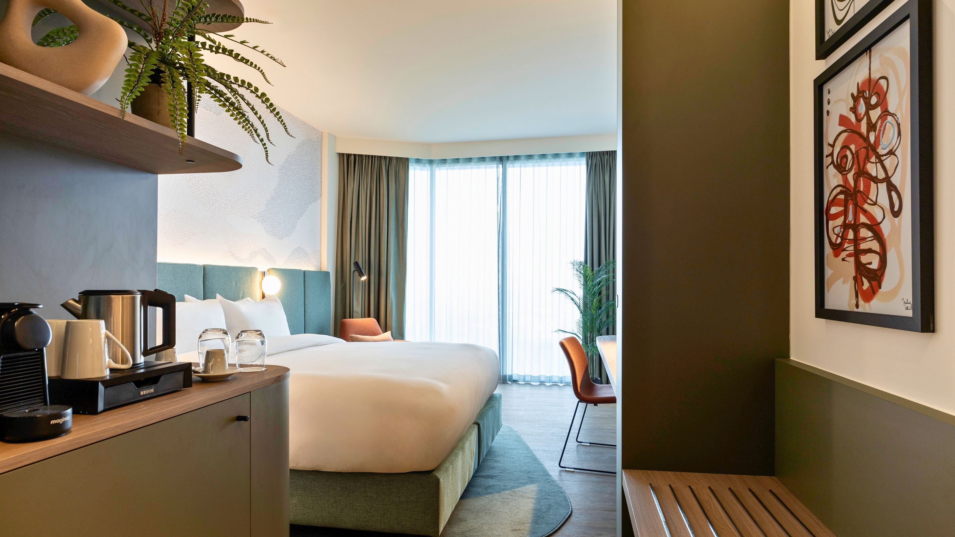 A modern hotel room with a neatly made bed, a coffee station, a desk, and a large window with curtains.