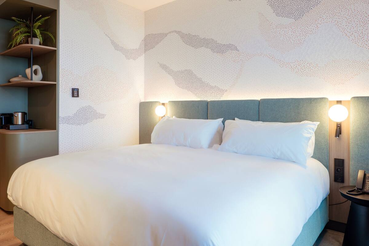 Ein ordentlich bezogenes Bett mit weißen Leinen, ein Kopfteil mit integrierter Beleuchtung und ein an der Wand befestigtes Telefon.