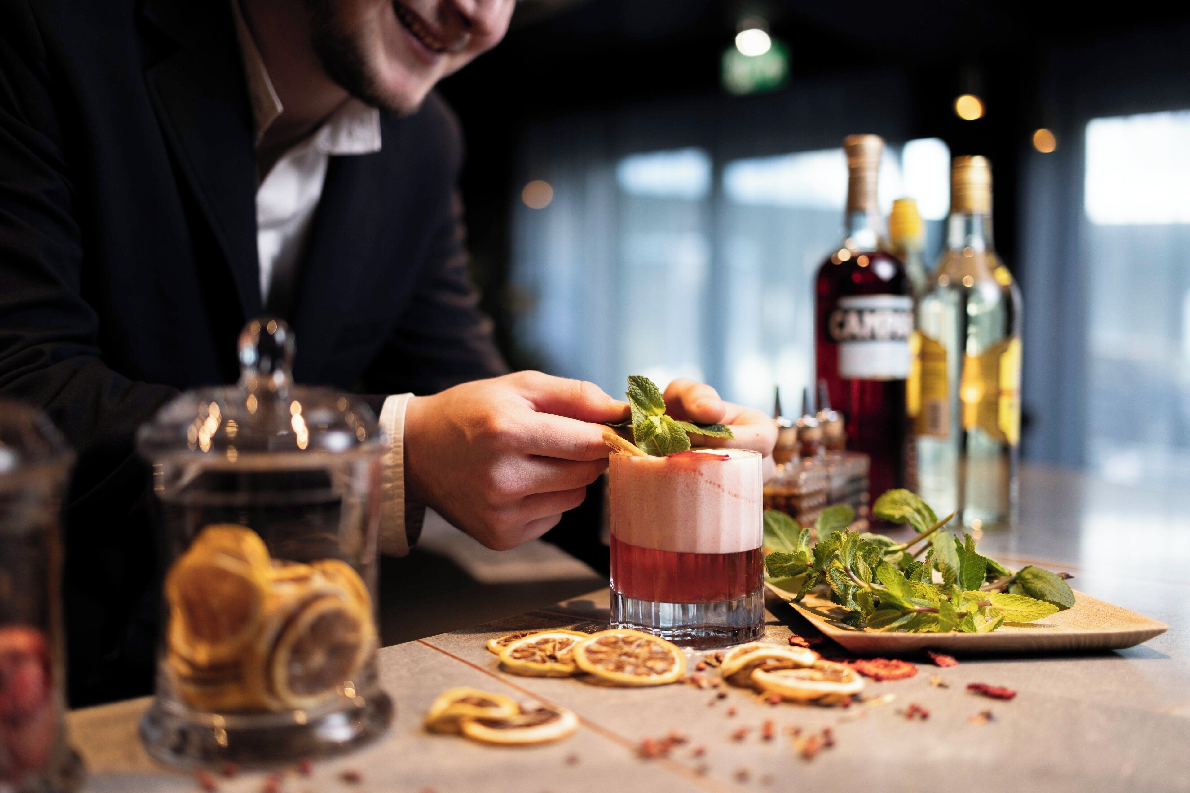 En person laver en drink med pynt, omgivet af forskellige flasker og pynt på et bord.
