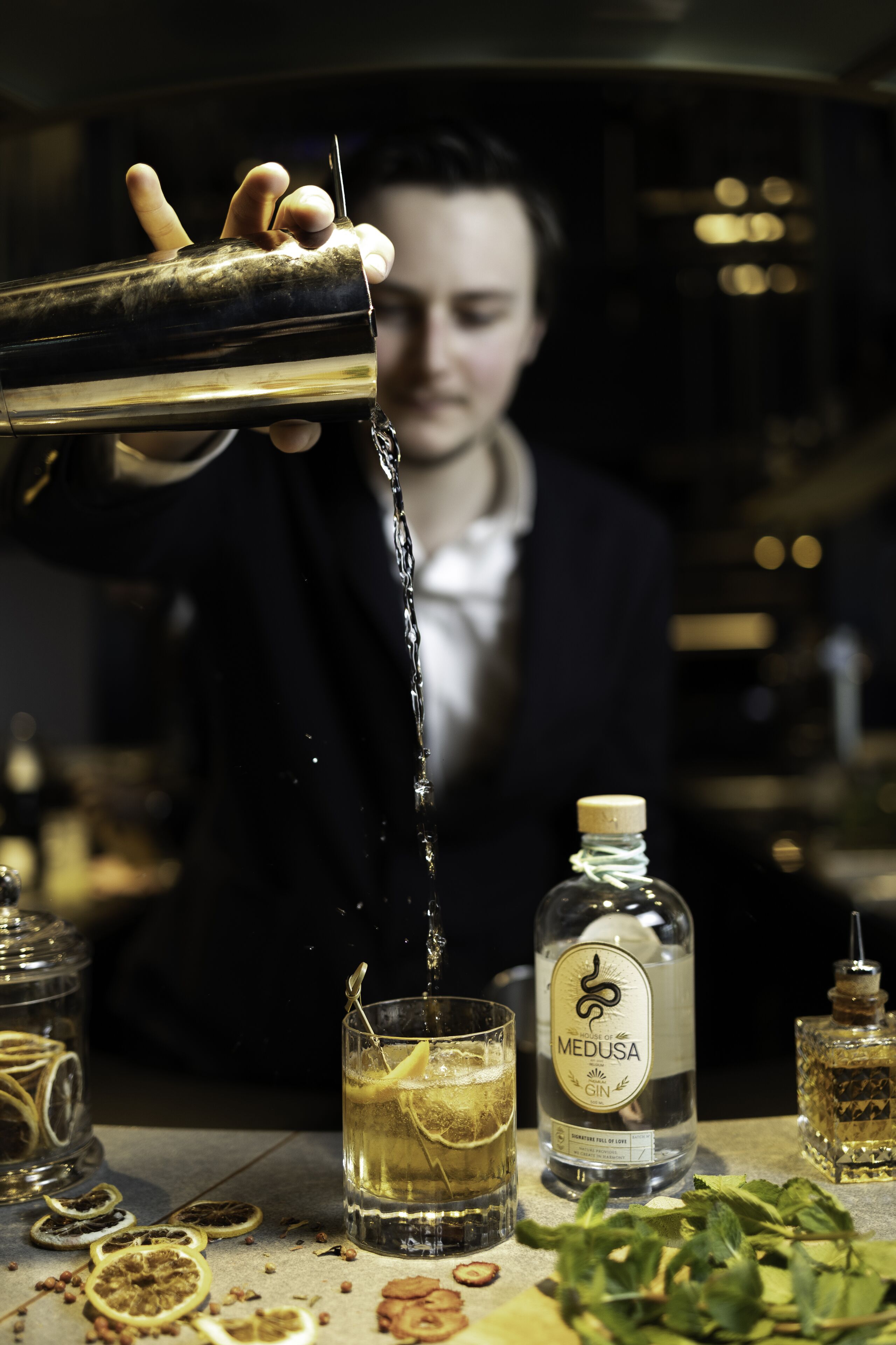 A bartender pouring a drink into a glass with a lemon slice, surrounded by various bottles and garnishes.