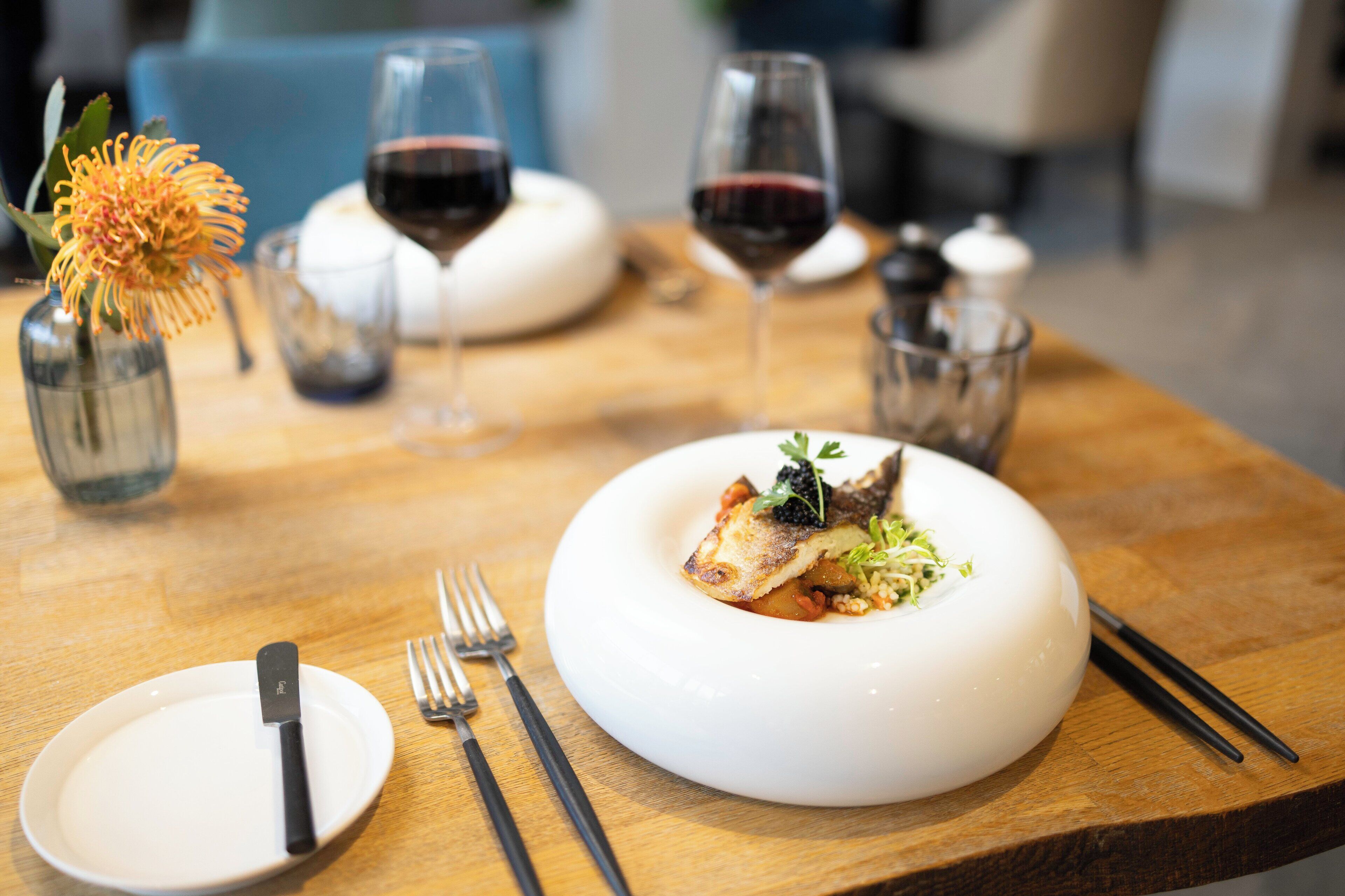 A dining table with a white plate containing a fish dish, accompanied by a fork, knife, and chopsticks. There are two glasses of red wine and a vase with a flower on the table.