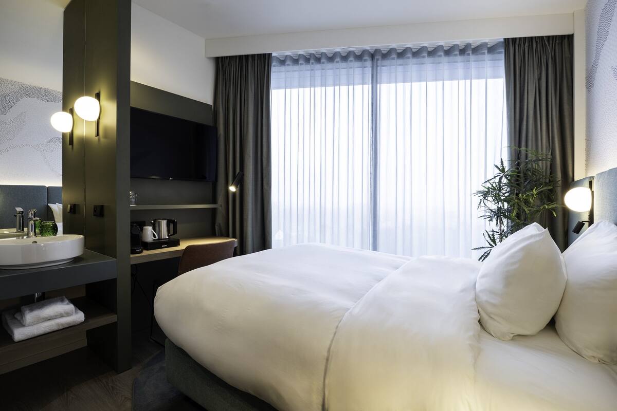 Ein Hotelzimmer mit einem großen Bett, einem Nachttisch mit Lampe, einem an der Wand befestigten Fernseher, einem Waschbecken und einem Fenster mit Vorhängen.