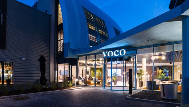 Una entrada de hotel moderna con fachada de vidrio, una recepción y un área de descanso con plantas.