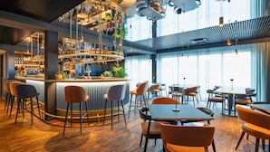 Un bar moderno con una barra curva, taburetes, y una variedad de botellas y vasos. Hay mesas y sillas para comer, y ventanas amplias que dejan entrar la luz natural.