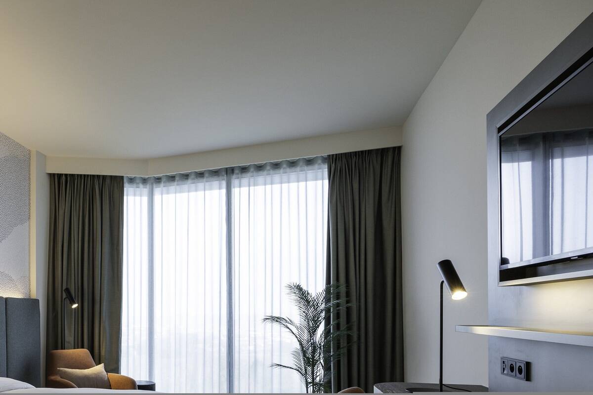 Ein modernes Hotelzimmer mit einem großen Bett, einer Sitzecke mit einem Sessel, einer Topfpflanze und einem großen Fenster mit Vorhängen.