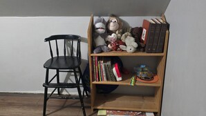 Una esquina de una habitación con una estantería de madera llena de libros, un peluche y una pequeña figurina. Una silla negra está colocada contra la pared.