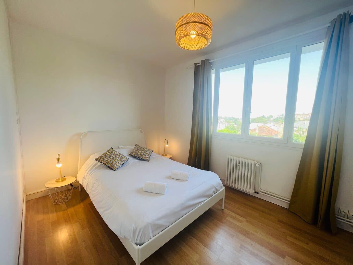 Een slaapkamer met een wit bed, twee bedkussens met een patroon, een bedlampje, een hanglamp, een raam met gordijnen, en een uitzicht op gebouwen.