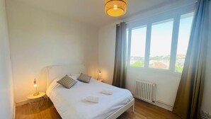 Un dormitorio con cama blanca, dos almohadones estampados, una lámpara de noche, un aplique, una ventana con cortinas y vistas a edificios.