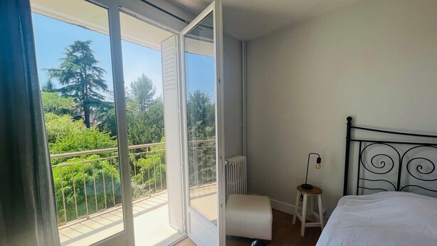 Un dormitorio con una cama, un taburete pequeño y vistas a la vegetación a través de un ventanal corredero.