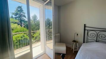 Une chambre à coucher avec un lit, un petit tabouret et une vue sur la verdure grâce à une porte-fenêtre coulissante.