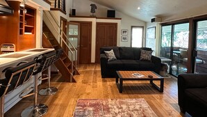 Un salón espacioso con piso de madera, un sofá negro, una mesa de centro y una escalera que lleva a un nivel superior.