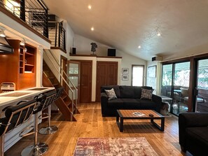 A spacious living area with a wooden floor, a black sofa, a coffee table, and a staircase leading to an upper level. - Aspen Downtown with Patio, Fireplace, Parking, W/D (Aspen)