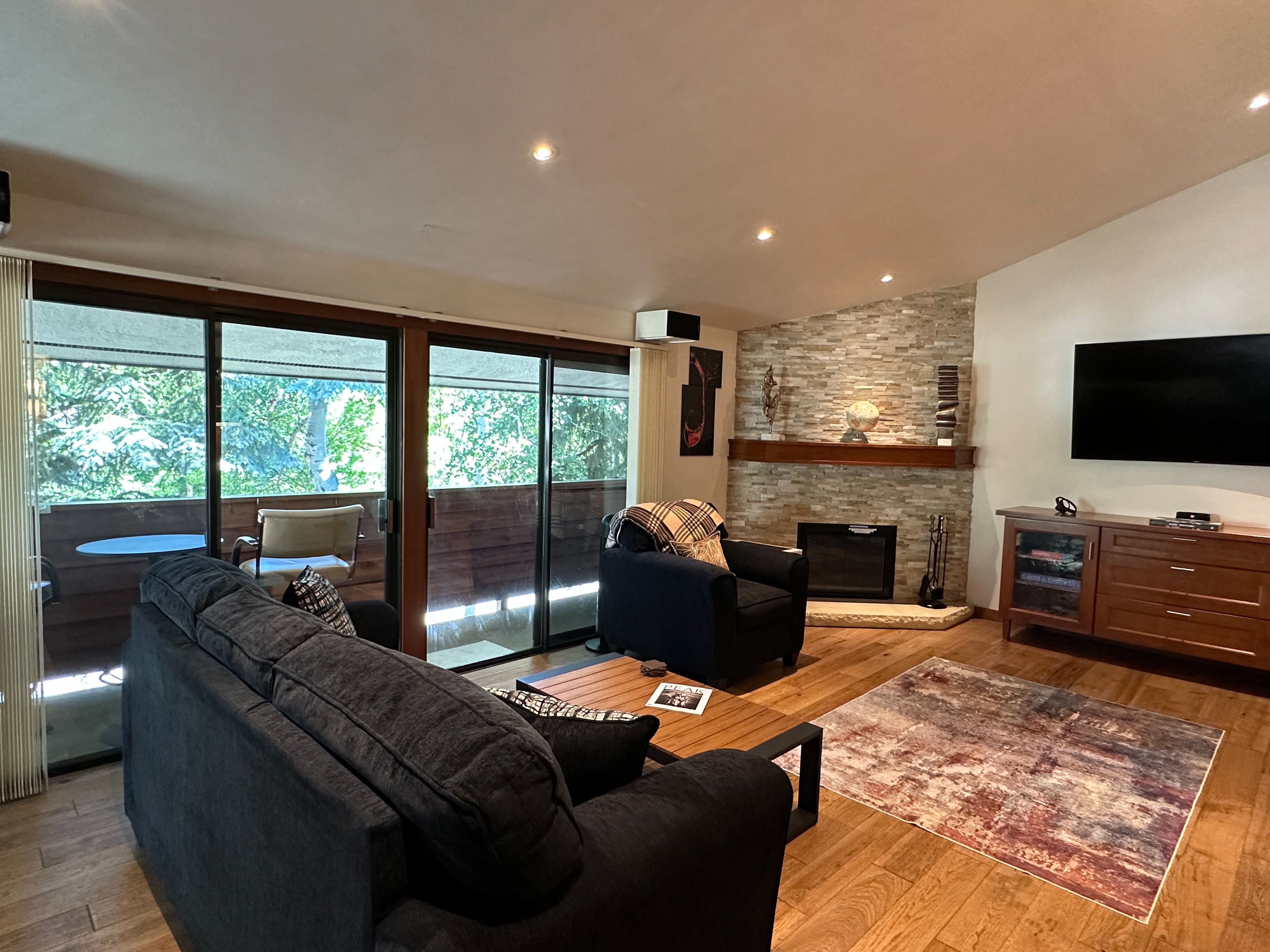 A modern living room with a large sofa, a coffee table, and a fireplace. There is a television mounted on the wall and a balcony with a table and chairs.