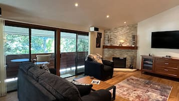 A modern living room with a large sofa, a coffee table, and a fireplace. There is a television mounted on the wall and a balcony with a table and chairs.