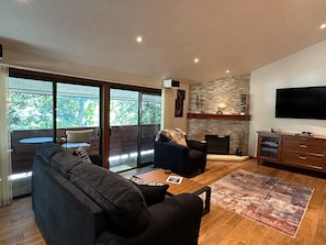A modern living room with a large sofa, a coffee table, and a fireplace. There is a television mounted on the wall and a balcony with a table and chairs.