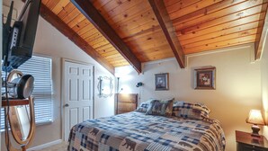 Een slaapkamer met een houten plafond, een bed met een quilt met een patroon, een dressoir en een lamp op een nachtkastje.