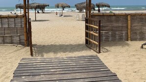 A wooden pier leading to a beach with thatched umbrellas and a bamboo fence.