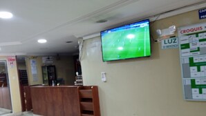 A reception area with a television mounted on the wall displaying a soccer game, a wooden reception desk, and a bulletin board with various notices. - Hotel Residencial Miraflores (Loja)
