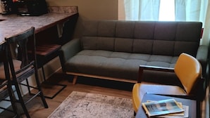 Ein Wohnzimmer mit einer Couch, zwei Sesseln, einem Couchtisch und einer Küchenzeile mit Schränken und einer Mikrowelle.